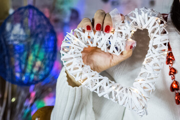 Closeup picture. Woman in white sweater holds in hand wooden heart with electric garland. New year decoration. Fingers with red nails. Christmas clothes. Tender and love concept. Saint Valentains day 