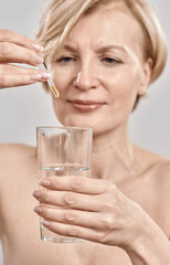 Close up of hand of attractive middle aged woman holding dropper with medication and glass isolated over grey background