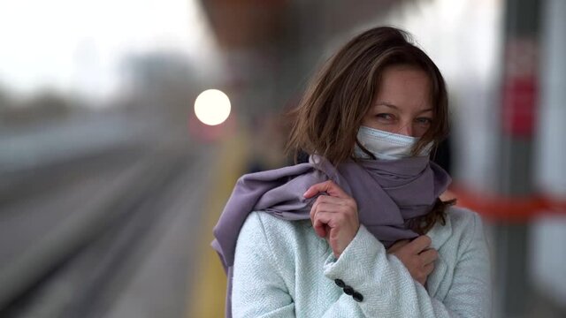 woman with facial mask from respiratory infection and scarf is standing on railway platform at autumn day