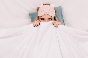 Adorable caucasian girl in sleep mask posing in bed. Interested woman hiding under blanket.