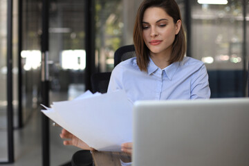 Attractive cheerful business woman checking paper documents in office, working on laptop.