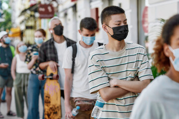 Fototapeta na wymiar Young asian guy wearing mask waiting, standing in line with other people, respecting social distancing to collect his takeaway order from the pickup point during coronavirus lockdown