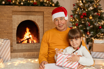 Fototapeta na wymiar Grandfather with her granddaughter sitting in living room and looking at camera, child holding present box, posing on floor on soft carpet near fir tree and fireplace.