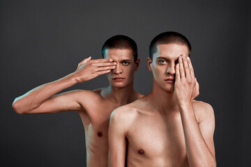 Studio shot of two young caucasian half naked twin brothers covering one eye with hand and looking at camera while posing together in studio