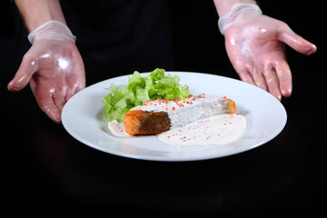 Obraz na płótnie Canvas The cook serves a prepared fish dish. Salmon in creamy caviar sauce on a white plate. Lettuce leaves. Photo on a black background. Unrecognizable person.