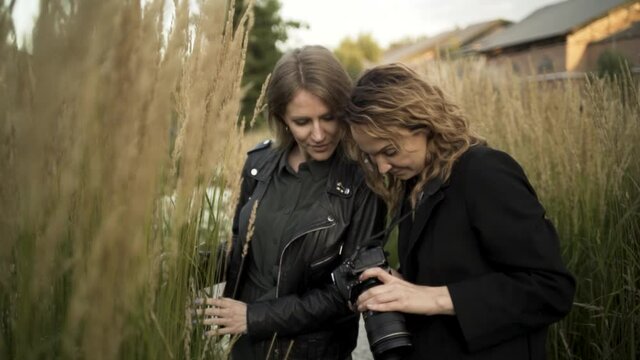 Woman photographer showing shots to a model while standing in a field with long grass. Action. Two women in wheatgrass looking at the professional camera, details of a photosession.
