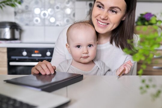 Portrait Of Mom And Baby Son Sitting At Home In Kitchen Looking In Laptop Screen