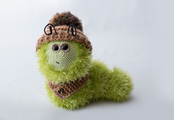 Green, fluffy, funny knitted caterpillar toy sitting on a white background. She is wearing a warm hat with a pompom and a small button-down scarf. Image with selective focus.