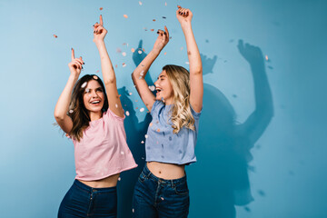 Cute girls dancing with hands up. Studio shot of carefree female friends fooling around on blue...