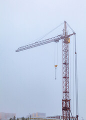 Tower industrial construction crane in the fog. Gray blue background. The roofs of houses are visible below. Vertical photography
