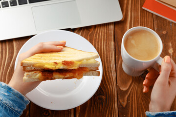 Hands of a woman holding sandwich above working table with laptop