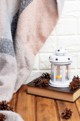 Christmas background with white candle lantern on old brown book with forest cones and warm scarf. Brick wall, wood background. Vertical orientation.