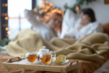 Two cups of tea, a saucer with lemon and a teapot stand on a wooden tray by the bed against the silhouettes of people lying in bed. Image with selective focus.