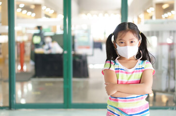 Asian child or kid girl smile wearing N95 white face mask for close mouth nose and flu cough sick and protect PM2.5 dust or influenza covid-19 and coronavirus to arms folded on department store door