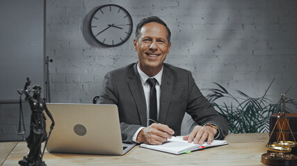  insurance agent looking at camera near notebook, laptop and statuette of justice on table