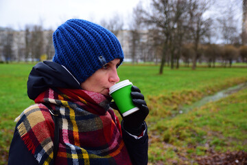 An adult woman drinks coffee while walking in the park in late autumn.