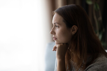 Pensive young woman looking in distance at window, thinking or life problems, suffering from loneliness or depressive mood, regretting mistakes, having psychological mental problems, copy space.