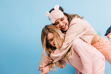 Gorgeous girl in sleep mask embracing friend. Studio shot of sisters in pajamas expressing positive emotions.