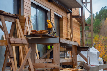 Stationary circular saw on the background of a house under construction. Installation for the preparation of timber beams. Concept - facing the house with wood. Services for facing cottages.