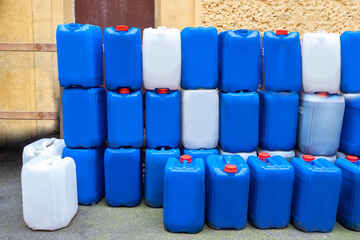 White and blue water-cans stand on top of each other. Lots plastic cans on background on wall. Water-can for water under open sky. Plastic tanks for transporting water. Plastic containers for liquid