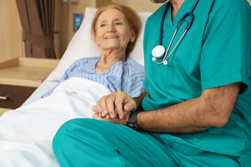 Doctor or assistance staff talking with an elder patient woman and hold the hand while sitting on bed in the hospital, healthcare and encouragement concept