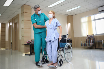 Fototapeta premium Doctor or assistance staff wear face mask and help or assist an elder patient woman while try to standing or walking or sitting on wheelchair in the hospital, eldercare concept
