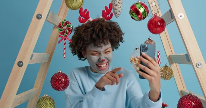 Funny woman makes face of christmas reindeer tries to scare or surprise friend by video call holds mobile phone has online communication uses free internet connection foolishes around at home