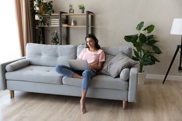 Fototapeta premium Full length focused young arabian indian woman wearing wireless headset with microphone sitting on sofa, holding distant online video call web camera meeting with colleagues, working from home.