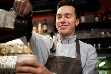 a staff member in a small restaurant works in an apron and shirt. Barista waiter prepares sells coffee Americano cappuccino with a takeaway in the cafe. pours a paper Cup from the teapot into a Cup