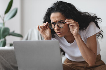 Young woman with bad eyesight using laptop, trying to work - obrazy, fototapety, plakaty