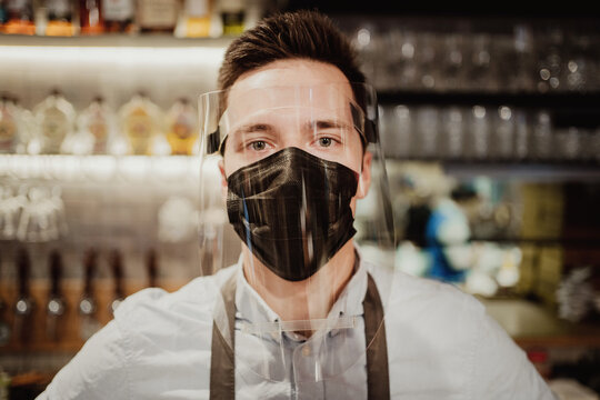 restaurant employee wearing a protective mask against the covid pandemic virus cafe bistro. portrait of a waiter a young man of dark Caucasian appearance stands near the bar. clothing apron and shirt.