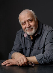 An adult gray-haired man in age, looking at the camera, his hands folded on the table. Portrait, studio, dark background, close-up, focus on the face.