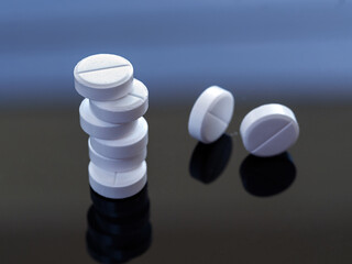 White round tablets are piled on top of each other. There are two pills next to the stack. Medical background with pills on a black - blue mirrored background close up. View from above.
