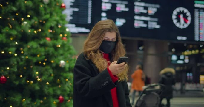 Woman in face mask scroll on phone application in christmas tree holiday setting at busy train station or airport arrival departure hall. New normal social distance travelling in 2020