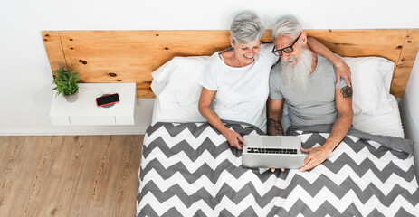 Trendy senior couple sitting in bed having video call with laptop - Joyful elderly lifestyle and technology concept - Focus on faces