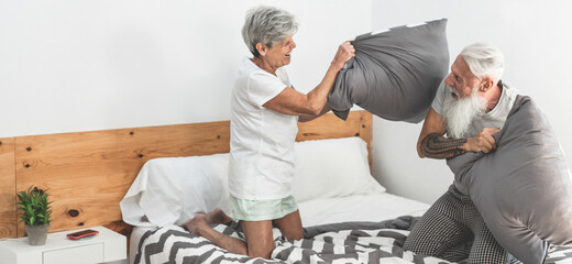 Senior couple doing pillow battle inside home bedroom - Happy mature people enjoying time together...