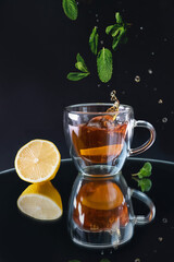 a glass cup with black tea, a splash in the cup from falling lemon and falling mint leaves with reflection on black background, lemon and mint leaf arranged on the table - 396093790