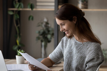 Smiling millennial european woman looking through paper letter correspondence, reading good news...