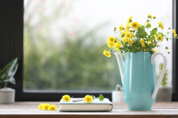Yellow Chrysanthemum in teal teapot vase with notebook and plant pot in front of windows 
