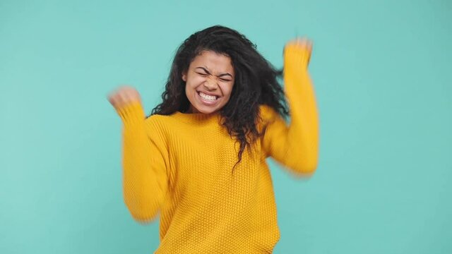 Surprised overjoyed young african american woman in yellow sweater isolated on blue background studio. People lifestyle concept. Doing winner gesture clenching fists applause cheering clapping hands