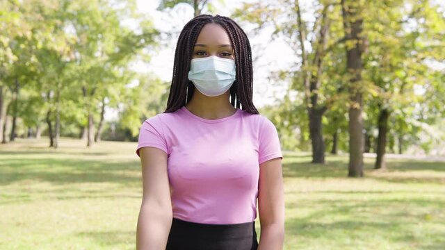 A young black woman in a face mask shows a thumb up to the camera and nods in a park on a sunny day