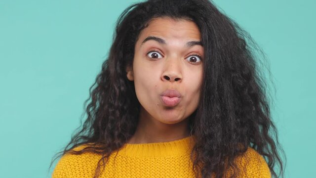 Close up of cheerful young african american woman in sweater isolated on blue turquoise background in studio. People lifestyle concept. Fooling around showing making fish face lips put hands on cheeks