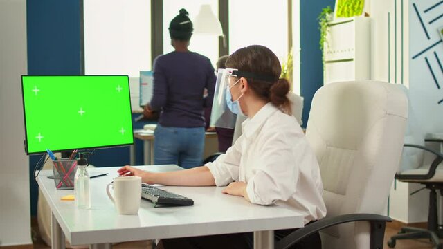 Company manager with face mask typing on computer with green screen in new normal business office respecting social distance during global pandemic. Woman Looking at mockup, copy space, chroma display