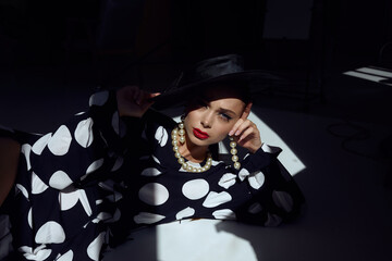 Woman with black hat wide and polka dot dress posing for fashion on a black background with shadow and rays of light on face