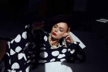 Woman with black hat wide and polka dot dress posing for fashion on a black background with shadow and rays of light on face - 396098986
