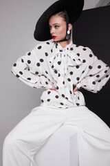 Woman with black hat wide white trousers and polka dot free shirt posing for fashion on a white and black background