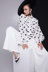 Woman with black hat wide white trousers and polka dot free shirt posing for fashion on a white and black background