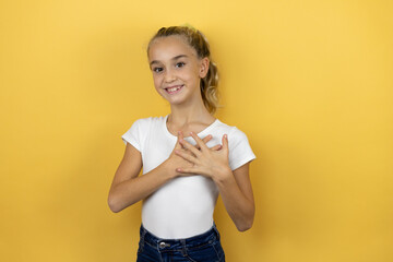 Young beautiful child girl standing over isolated yellow background smiling with her hands on her chest and grateful gesture on her face.