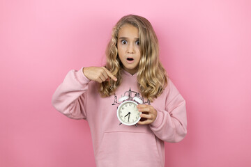 Young beautiful child girl standing over isolated pink background serious holding and pointing a clock