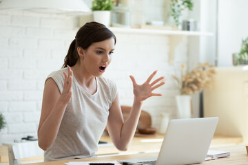 Angry shocked young woman looking at laptop screen, sitting at table in modern kitchen at home, stressed amazed female with open mouth reading bad news in email or message, financial problem